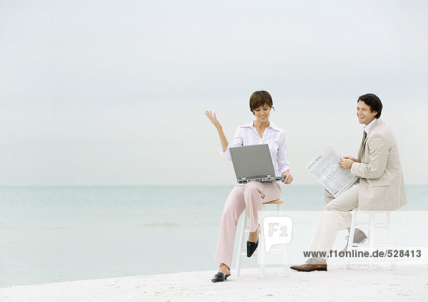 Zwei Geschäftsleute sitzen auf Hockern am Strand  einer mit Laptop  der andere mit Zeitung lesen