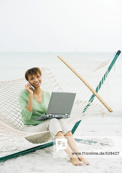 Frau sitzt auf Hängematte am Strand  mit Handy und Laptop