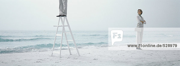 Woman standing on beach with arms crossed  looking toward man standing on ladder