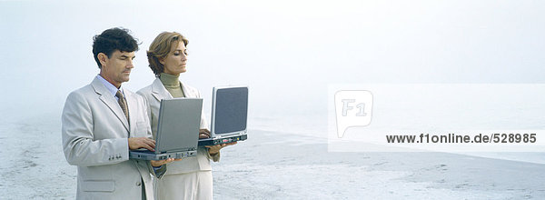 Geschäftsmann und Frau stehend  Laptops am Strand haltend