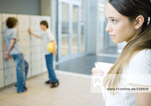 Teenage girl holding notebook  looking away  two teen boys standing near lockers in background