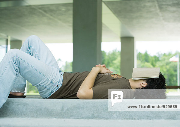 Male college student lying on ground with book covering face