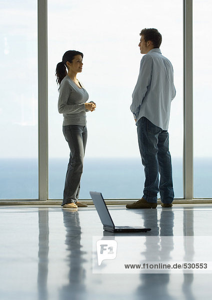 Junger Mann und Frau stehen von Angesicht zu Angesicht am Fenster  Laptop auf dem Boden  volle Länge