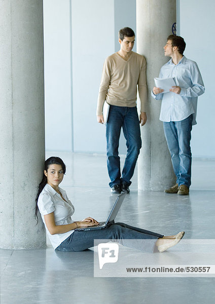 Junge Frau auf dem Boden sitzend  mit Laptop  zwei Männer hinter ihr stehend
