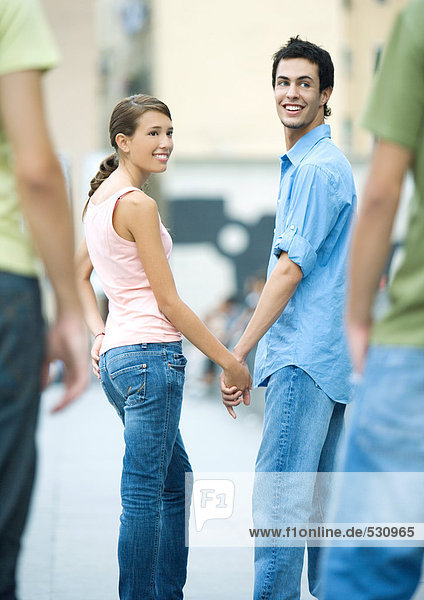 Teenager-Paar hält sich an den Händen,  schaut über die Schultern zu Freunden
