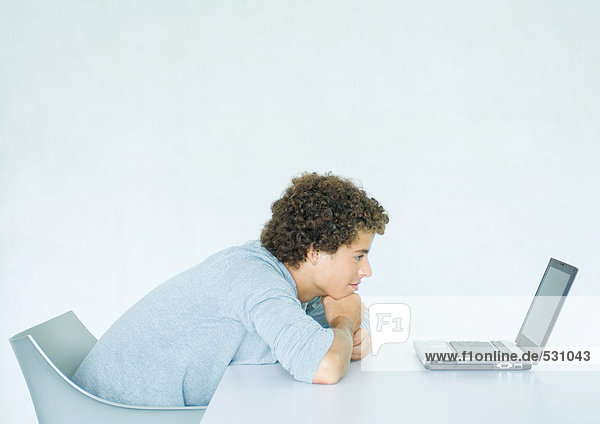 Junger Mann sitzend  auf dem Schreibtisch lehnend  mit Blick auf den Computer