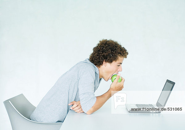 Junger Mann sitzt am Tisch  isst Apfel und benutzt Laptop