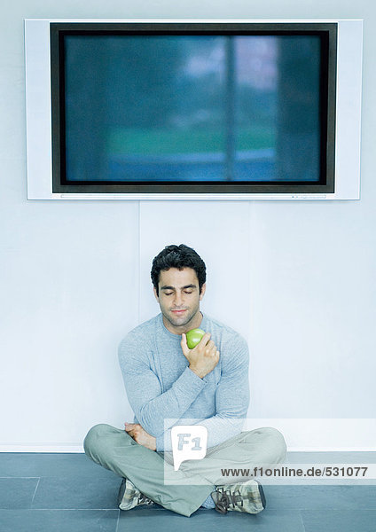 Mann sitzt auf dem Boden  isst Apfel  Breitbild-Fernseher an der Wand über Kopf