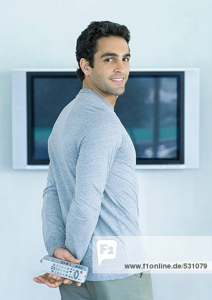 Mann steht vor einem Breitbild-Fernseher  hält zwei Fernbedienungen und lächelt über die Schulter.