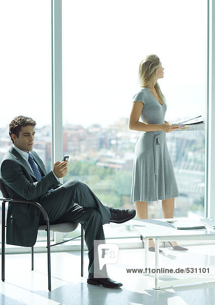 Geschäftsmann und Frau warten in der Lobby  Mann überprüft Handy  Frau steht im Hintergrund  hält Magazin