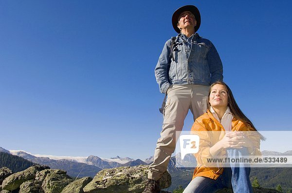Paar in den Bergen  Mann stehend  Frau auf Felsen sitzend