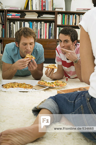 Three young people lying on floor  eating pizza