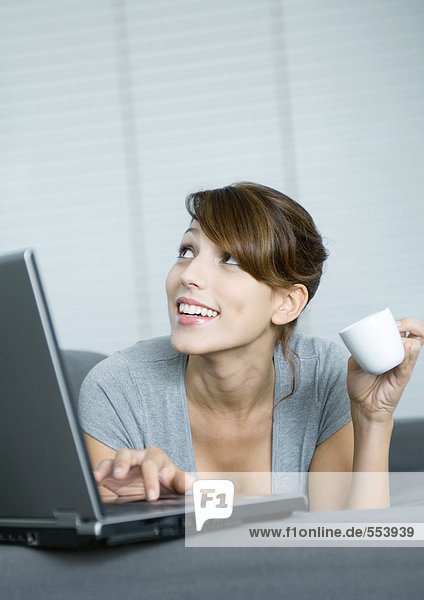 Young woman on couch  using laptop and holding coffee cup  looking up