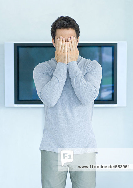 Mann steht vor dem Breitbildfernseher  Hände bedecken Gesicht