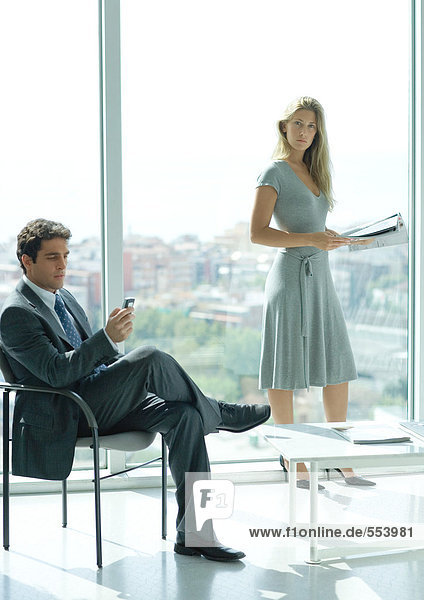 Bürolobby  Mann sitzt und schaut auf das Handy  während die Frau im Hintergrund steht und das Magazin hält.