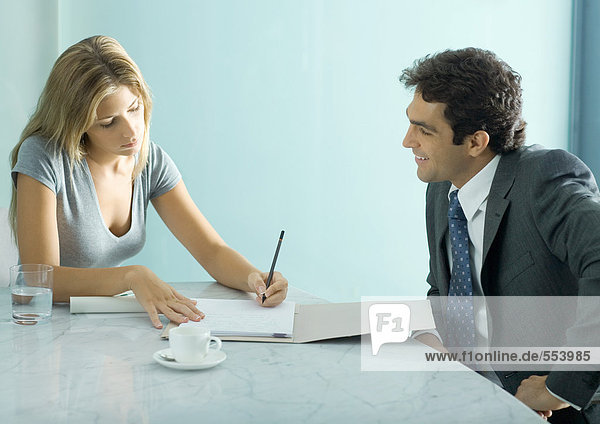 Geschäftsmann und Kollegin am Tisch sitzend  Frau beim Notieren