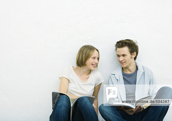 Teenagermädchen und junger Mann sitzen mit Büchern auf dem Boden und lächeln sich an.