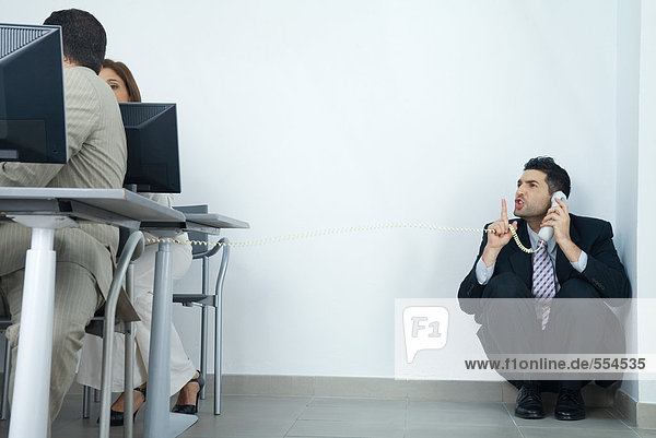 Businessman sitting in corner  holding telephone  looking at colleagues and holding finger in front of lips