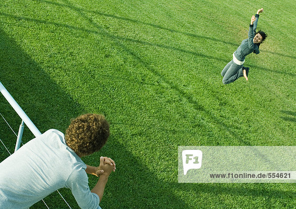 Teenager-Junge  der sich auf das Geländer stützt und das Teenager-Mädchen beim Springen auf Gras beobachtet.