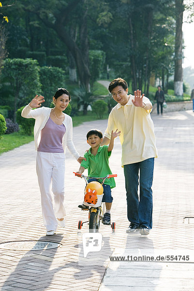 Boy riding bicycle  parents on either side  waving at camera