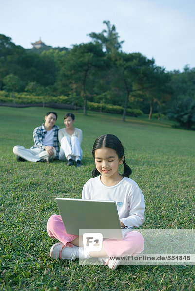 Mädchen sitzend im Gras mit Laptop  Eltern sitzend im Hintergrund