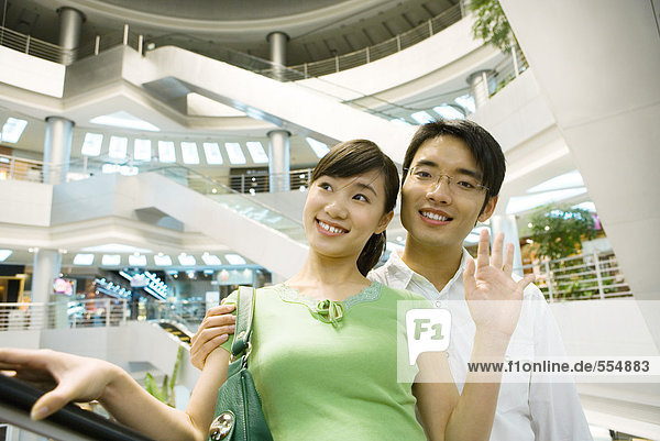 Couple taking escalator in shopping mall  woman waving