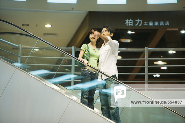 Couple taking escalator in shopping mall  man pointing out of frame