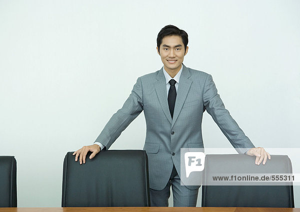 Geschäftsmann stehend mit Händen auf Stuhllehnen  lächelnd vor der Kamera  dreiviertel Länge