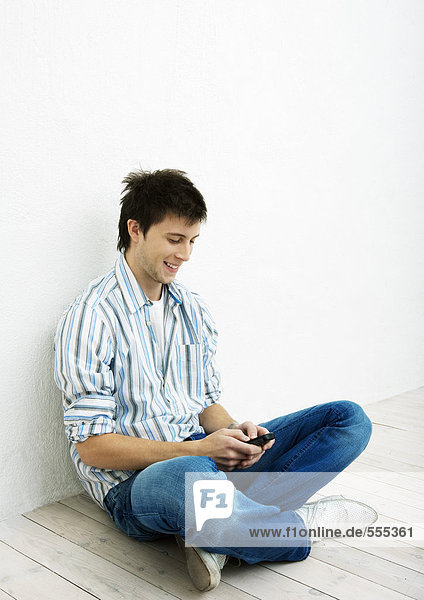 Junger Mann  der auf dem Boden sitzt und ein Handy benutzt.