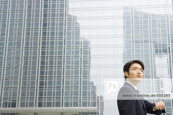 Junger Geschäftsmann hält das Handgelenk hoch  um die Zeit zu überprüfen  schaut weg  Bürogebäude im Hintergrund