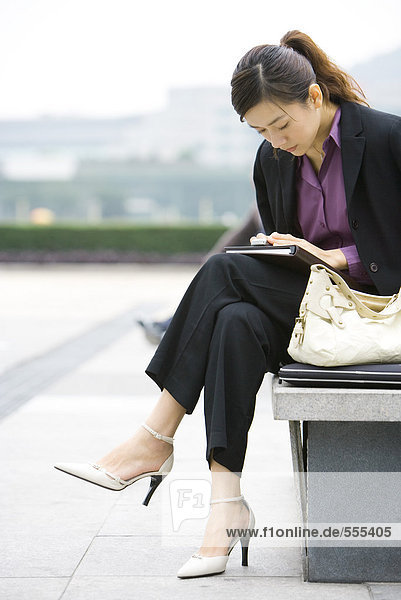 Junge Geschäftsfrau sitzt auf der Bank und schaut auf das Handy.