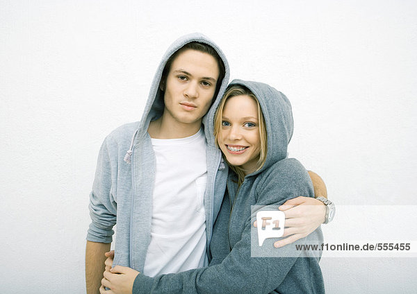 Junges Paar steht mit Armen umeinander  trägt Kapuzensweatshirts  schaut in die Kamera.