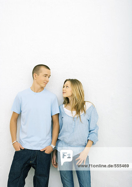 Young couple standing side by side  leaning against wall  looking at each other  three quarter length