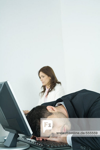 Businessman sleeping on keyboard while colleague works in background