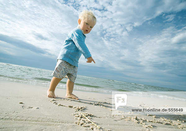 Kleinkind steht am Strand  schaut in die Kamera und zeigt auf Sand.