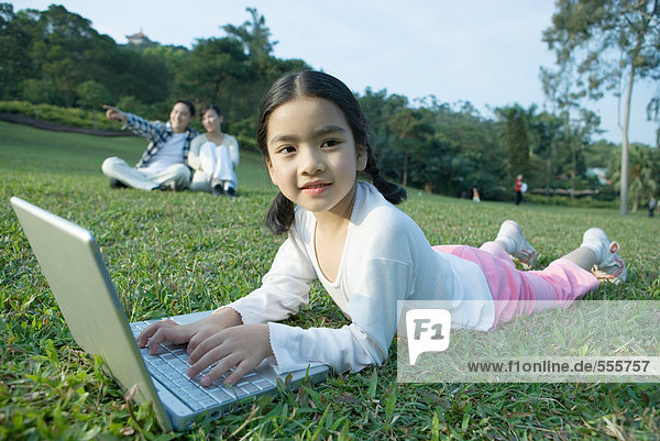 Mädchen im Gras liegend mit Laptop  Eltern im Hintergrund