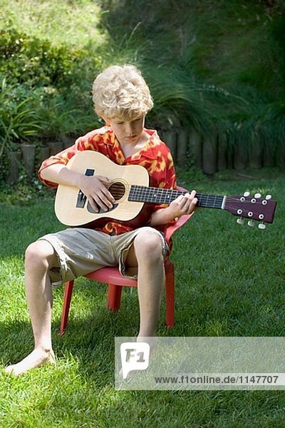 Young blond haired boy in bright red and yellow shirt playing a guitar outdoors.