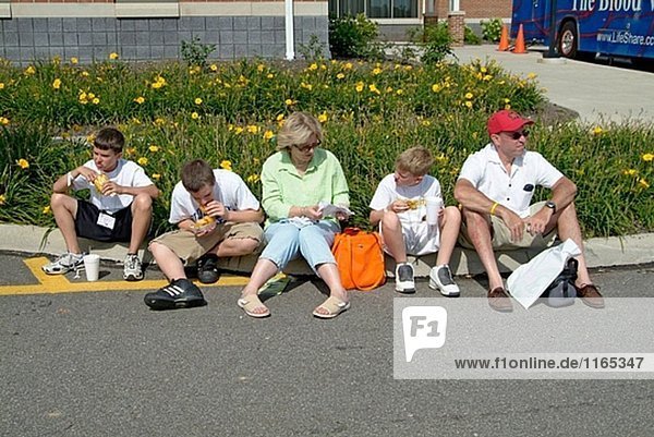 Familie entspannt auf den Bordstein einer Straße essen Mittagessen während des Besuchs einer festival