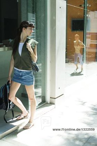 Young woman standing by entrance  looking over shoulder at friend arriving  reflected in window
