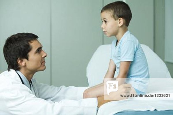 Junge sitzt auf dem Untersuchungstisch  Arzt schaut auf den Jungen  Profil