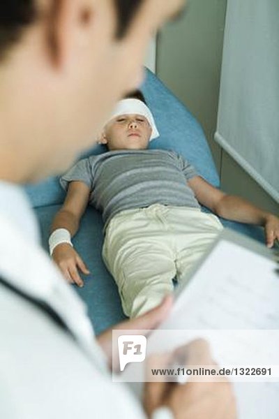Junge auf Untersuchungstisch liegend mit Stirnbandage und Arm  Arztbrief auf Klemmbrett
