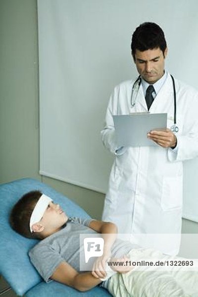 Junge auf Untersuchungstisch liegend mit Stirnbandage  Haltearm  Arzt-Schriftzug auf Klemmbrett