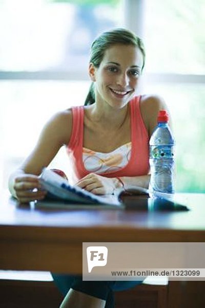 Junge Frau in Trainingskleidung  sitzend mit Magazin und Wasserflasche  lächelnd vor der Kamera