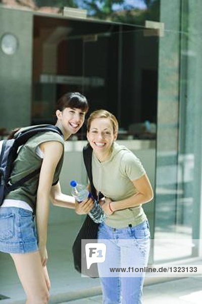 Zwei junge Frauen stehen draußen in Straßenkleidung und lächeln in die Kamera.