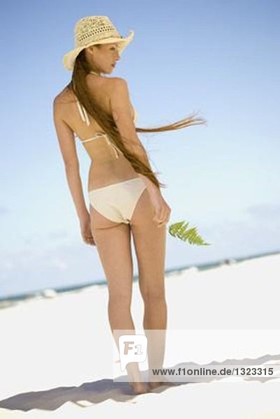 Frau mit Bikini und Sonnenhut am Strand  Blatt in der Hand  volle Länge  Rückansicht