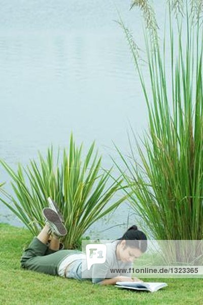 Junge Frau  die auf Gras in der Nähe eines Gewässers liegt und liest.