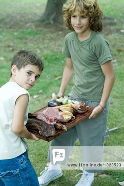 Zwei Jungen tragen ein Tablett mit gegrilltem und rohem Fleisch.