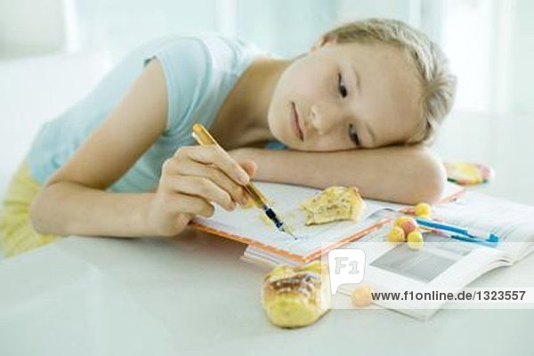 Mädchen am Tisch sitzend  Hausaufgaben machend  Kopf auf dem Arm liegend  kritzelnd  Essen auf Büchern verstreut