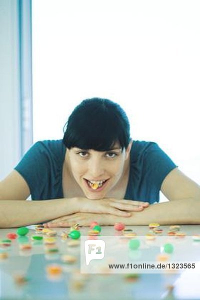 Junge Frau am Tisch sitzend  mit Süßigkeiten bedeckt  hält ein Stück Süßigkeiten zwischen den Zähnen.