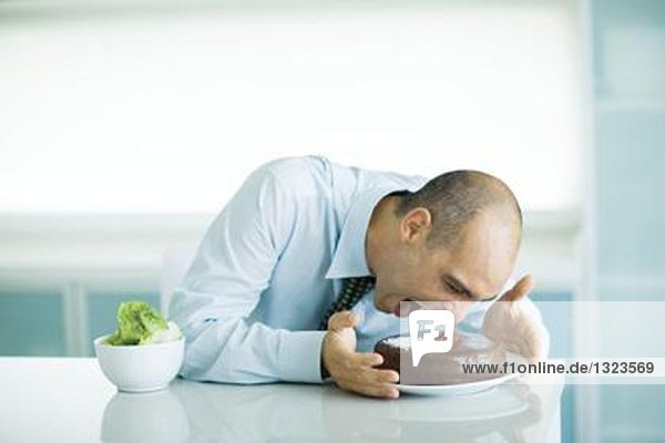 Mann am Tisch sitzend mit Schokoladenkuchen und Salatschüssel  zum Kuchen geneigt mit weit geöffnetem Mund